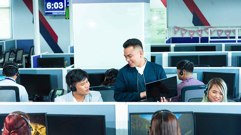 An image depicting agents in a customer support center. One is standing and holding a laptop while talking to another agent who is seated at his desk.